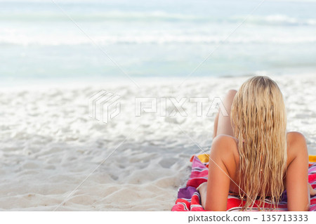 Adult female lying on stomach on striped towel at shore, wearing two-piece swimsuit, copy space Adult female lying on stomach on striped towel at shore, wearing two-piece swimsuit, copy space 135713333
