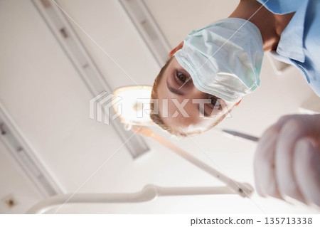 Female dentist wearing mask holding probe in clinic under overhead lamp and ceiling panels Female dentist wearing mask holding probe in clinic under overhead lamp and ceiling panels 135713338