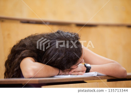 female student resting head on arms atop open notebook with digital wristwatch on desk in classroom 135713344