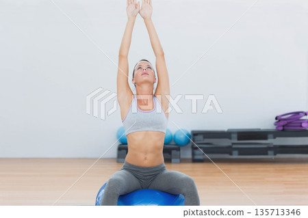 Female stretching arms upward sitting on blue stability ball in gym with balls, steps, purple mats 135713346