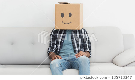 Adult male sitting on light grey sofa against white wall, wearing plaid shirt, cardboard box mask Adult male sitting on light grey sofa against white wall, wearing plaid shirt, cardboard box mask 135713347