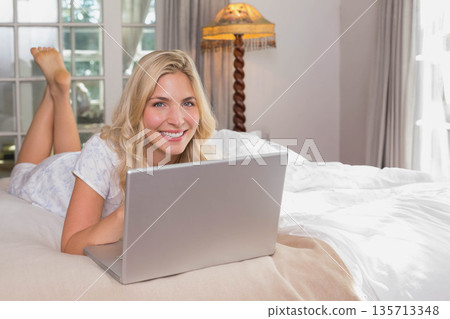 Blonde woman lying on stomach on white linens in bedroom working on laptop beside fringed lamp Blonde woman lying on stomach on white linens in bedroom working on laptop beside fringed lamp 135713348