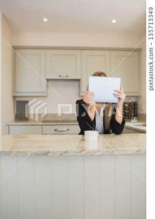 Businesswoman sitting at home behind kitchen island holding tablet computer obscuring face with mug 135713349