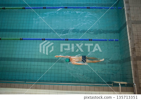 Male swimmer performing front crawl in tiled pool between lane ropes near ladder and drainage grate 135713351