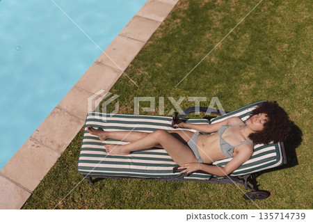 African American woman in twenties reclining on striped lounge chair poolside holding sunglasses African American woman in twenties reclining on striped lounge chair poolside holding sunglasses 135714739