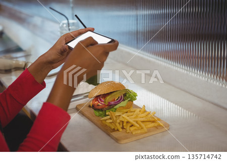 African American man in red shirt snapping smartphone picture of burger fries drink at cafe counter African American man in red shirt snapping smartphone picture of burger fries drink at cafe counter 135714742