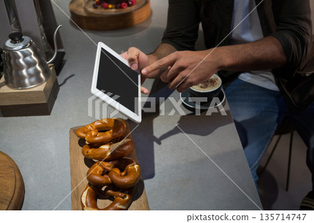 African American man wearing jacket tapping white tablet at cafe counter with pretzels cappuccino African American man wearing jacket tapping white tablet at cafe counter with pretzels cappuccino 135714747