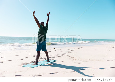 Senior African American man stretching on teal yoga mat on beach with footprints facing sea 135714752