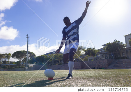 African American man wearing striped jersey kicking rugby ball on red tee on grass rugby pitch African American man wearing striped jersey kicking rugby ball on red tee on grass rugby pitch 135714753