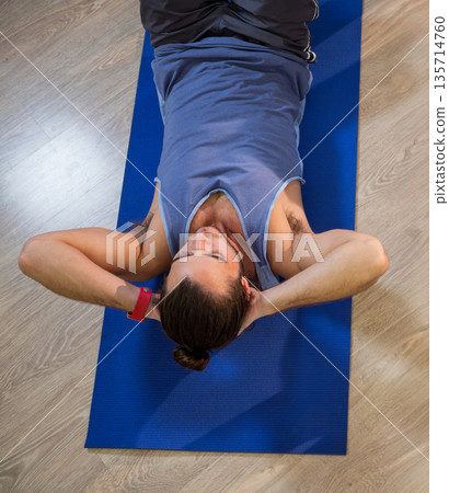 Man lying on mat performing crunch on exercise room floor wearing sleeveless top fitness tracker Man lying on mat performing crunch on exercise room floor wearing sleeveless top fitness tracker 135714760