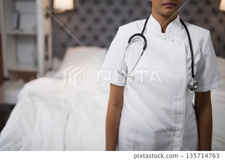 Female nurse wearing white uniform and stethoscope standing in bedroom by unmade bed with lit lamp 135714763