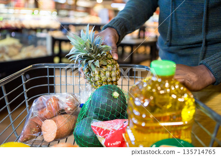 African American man placing pineapple into cart with oil bottle, powder bag in produce section 135714765