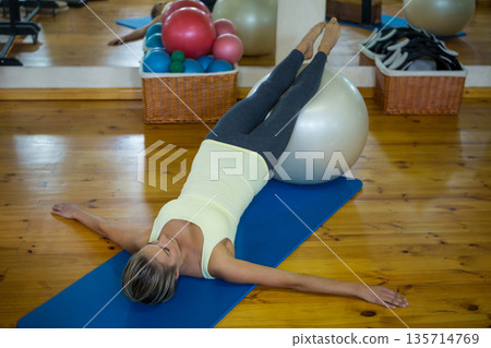 Mid adult woman in sportswear lying on mat raising legs onto silver ball in fitness studio Mid adult woman in sportswear lying on mat raising legs onto silver ball in fitness studio 135714769
