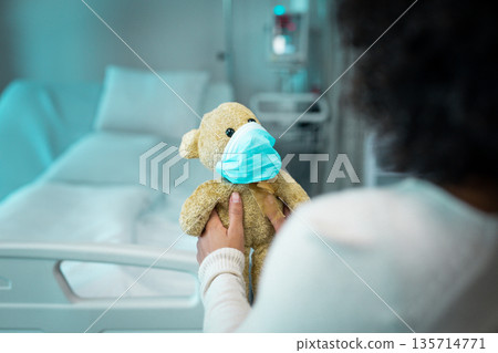African American woman holding teddy bear wearing blue mask with hospital bed monitor and IV stand African American woman holding teddy bear wearing blue mask with hospital bed monitor and IV stand 135714771