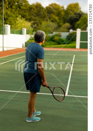 Senior male in sportswear standing facing tennis net on club court holding racket beside fence Senior male in sportswear standing facing tennis net on club court holding racket beside fence 135714780