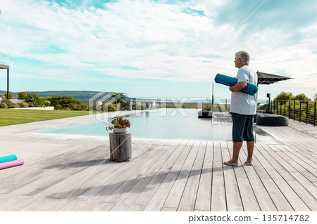 Senior man standing barefoot on wooden deck beside infinity pool holding teal yoga mat, copy space 135714782
