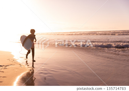 Mid-adult African American woman running on shore carrying white surfboard at sunrise, copy space Mid-adult African American woman running on shore carrying white surfboard at sunrise, copy space 135714783