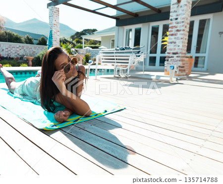 Woman lying face down on striped turquoise towel beside pool lifting sunglasses near patio chairs Woman lying face down on striped turquoise towel beside pool lifting sunglasses near patio chairs 135714788