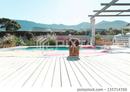 African American woman sunbathing wearing yellow bikini top on towel at pool deck with pool ring 135714789