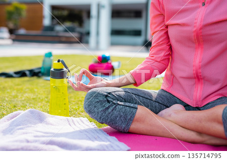 Woman meditating on yoga mat on lawn, forming mudra near towel and water bottles beside dumbbells Woman meditating on yoga mat on lawn, forming mudra near towel and water bottles beside dumbbells 135714795