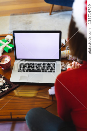 Asian woman wearing red sweater, Santa hat typing on laptop at home with mug, gift boxes 135714799