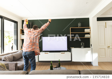 Middle-aged man celebrating raising arms in living room facing TV holding yellow drink can Middle-aged man celebrating raising arms in living room facing TV holding yellow drink can 135714810