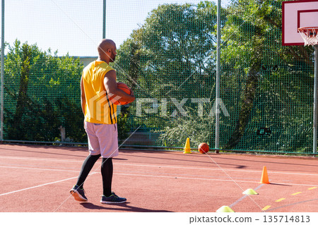 African American man in jersey holding basketball under arm near cones by hoop on red court African American man in jersey holding basketball under arm near cones by hoop on red court 135714813