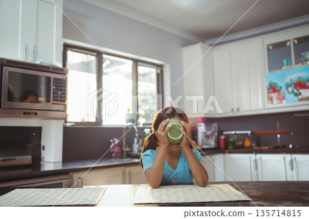 Green mug steaming on wooden table near window with potted plant, microwave on dark countertop 135714815