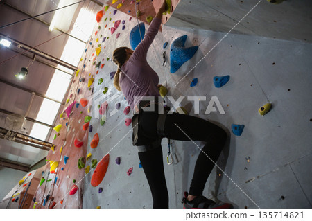 Female climber ascending climbing wall in gym with holds under lights and beams using harness, rope Female climber ascending climbing wall in gym with holds under lights and beams using harness, rope 135714821