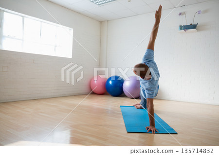 Man performing side plank on blue mat in yoga studio with pastel balls, plant, copy space 135714832