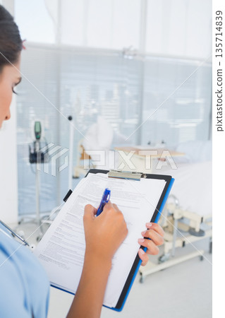 Adult female nurse standing in hospital room writing on clipboard beside wheeled bed, copy space 135714839