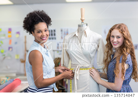 Female colleagues using measuring tape taking waist measurements on dress form in fashion studio 135714847