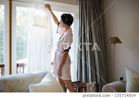 Woman wearing satin robe holding wedding dress on hanger in living area, copy space Woman wearing satin robe holding wedding dress on hanger in living area, copy space 135714854