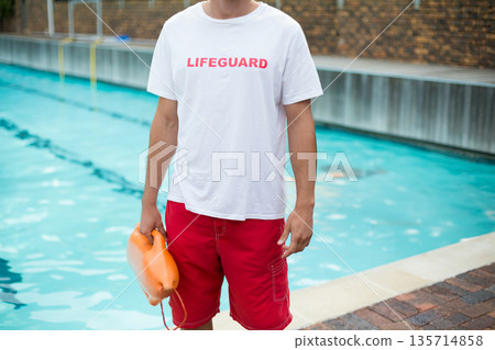 Lifeguard standing at pool edge holding orange rescue buoy beside turquoise water and brick deck 135714858