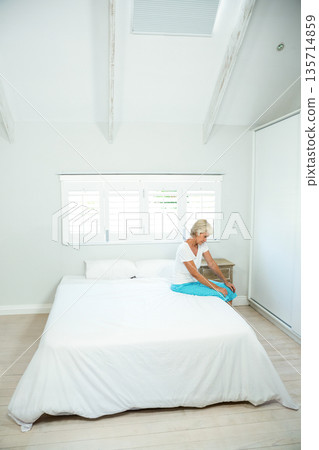 Senior woman sitting on right edge of bed under shuttered window in bedroom, copy space 135714859