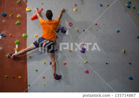 Male climber in orange tee ascending climbing wall with multicolored holds at gym with chalk bag 135714867