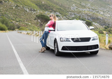Woman leaning against white sedan beside fence on mountain road wearing sunglasses, copy space Woman leaning against white sedan beside fence on mountain road wearing sunglasses, copy space 135714870