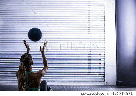 Woman wearing green tank top tossing black medicine ball on mat by shutter door in gym 135714871
