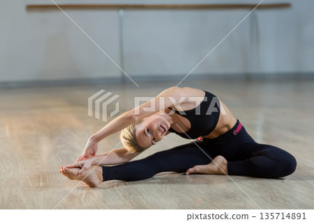 Female dancer in black sportswear performing side stretch on studio floor with wooden ballet barre 135714891