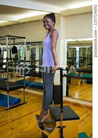 African American woman wearing purple activewear standing on pilates chair in studio rising on toes 135714892