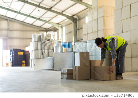 Female warehouse worker stacking boxes on warehouse floor with barrels and crates, copy space Female warehouse worker stacking boxes on warehouse floor with barrels and crates, copy space 135714903