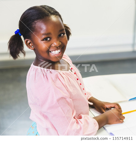 African American girl sitting at school desk writing in notebook with pencil and whiteboard behind 135714911