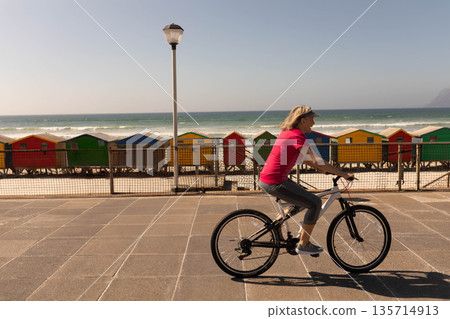 Senior woman riding bicycle along coastal path passing railing, colorful huts and lamp, copy space Senior woman riding bicycle along coastal path passing railing, colorful huts and lamp, copy space 135714913