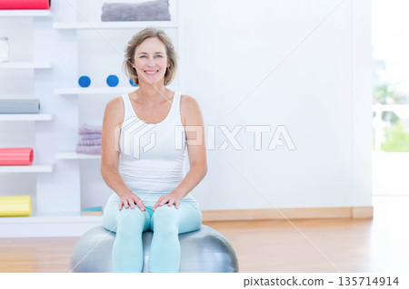 Woman balancing on exercise ball inside fitness studio with yoga mats and foam rollers, copy space 135714914