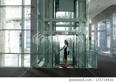 Female professional walking past glass elevator shaft inside corporate lobby with bag 135714916
