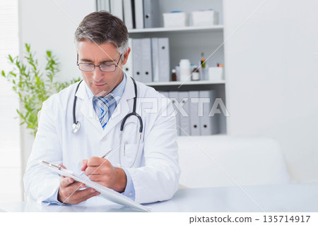 Male physician writing on clipboard with stethoscope at desk by potted plant in medical office 135714917