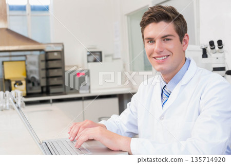 At lab under fume hood male researcher using laptop, microscope while smiling at camera, copy space At lab under fume hood male researcher using laptop, microscope while smiling at camera, copy space 135714920