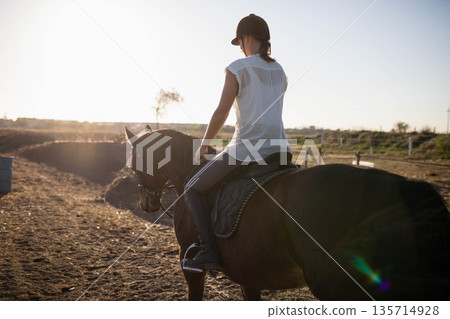 Female rider wearing brown helmet and holding reins sitting on saddled horse with bridle in paddock 135714928