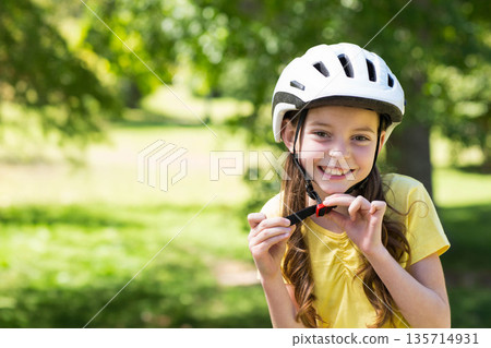 Female child fastening helmet chin strap standing in park wearing yellow tee, copy space Female child fastening helmet chin strap standing in park wearing yellow tee, copy space 135714931
