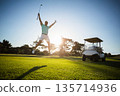 Male golfer jumping raising golf club wearing golf glove on fairway at sunset with golf cart 135714936